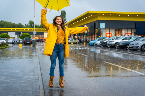 Young woman jumping in the water puddle. She is wearing a yellow raincoat and an umbrella.