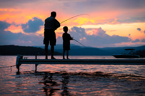 A grandfather is teaching his grandson to fish during summer. They are both standing on the dock. It is a beautiful summer day at sunset. Across the lake, there is a mountain, Quebec, Canada.