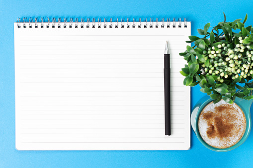 Blank notebook with a pen and a cup of coffee on a blue background, top view.