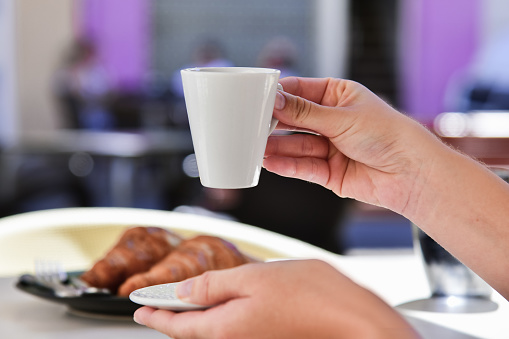 Close up of female hands holding a coffee cup and a plate with out of focus croissants in the back. Breakfast and lifestyle concept.