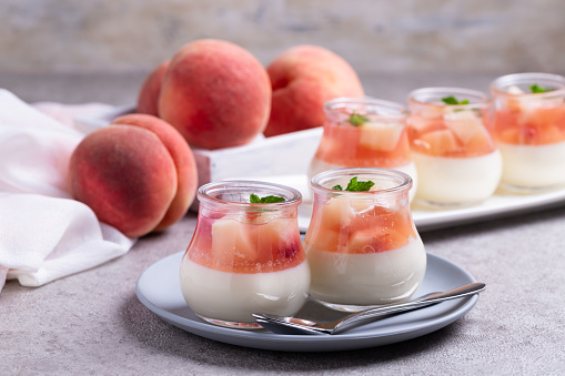 homemade panna cotta with slices of peach and peach jelly in glass jars on a gray concrete background.