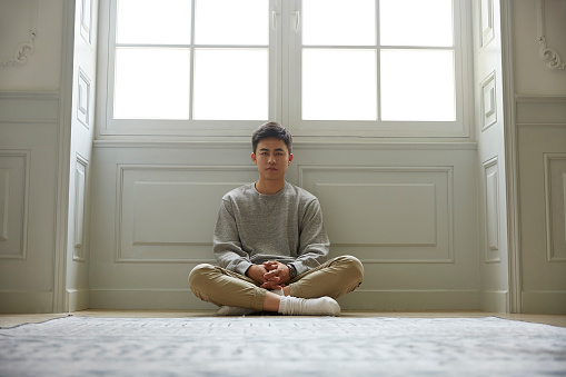young asian man sitting on floor at home legs crossed looking at camera
