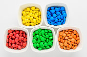Five squared bowls with small red, blue, green and orange coated chocolate candies similar to m&ms in a squared bowl isolated on white background, top view