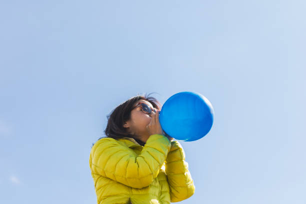 Asian woman wearing sunglasses blowing up a toy ballon outdoors.