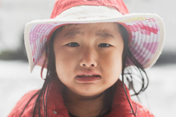 An adorable little girl stands outside on a cold winter day. She is wearing a hat with a frigid look on her face. Her eyes evoke sadness.