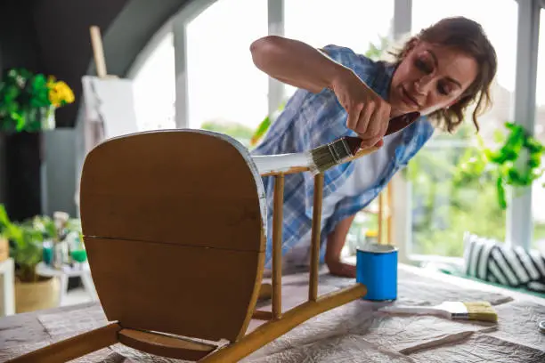 Wide shot of active mature woman working on a table and restoring her old chair with white paint.
