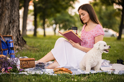 Side view of focused young woman reading an interesting book and drinking coffee while having a picnic with her cute dog on a public park.