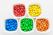 Five squared bowls with small red, yellow, blue, green and orange coated chocolate candies similar to m&ms in a squared bowl isolated on white background, top view