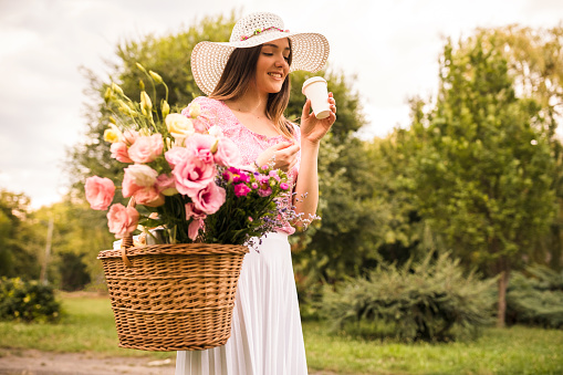Three quarter length of young woman walking in a natural area, wearing a summer hat and carrying a picnic basket with pink flowers while drinking coffee.