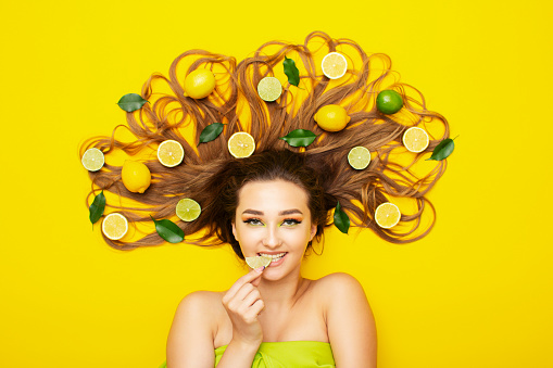 portrait of girl lying on yellow background with citrus fruits on long hair,young woman bites lemon wedge, food taste, energy