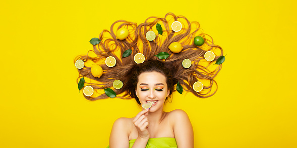 portrait of girl lying on yellow background with citrus fruits on long hair,young woman bites lemon wedge, food taste, energy