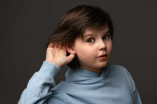 Curious young schoolboy 10-12 years old, holding hand at his ear, trying to overhear private conversation, dressed in casual clothes, posing at gray studio background