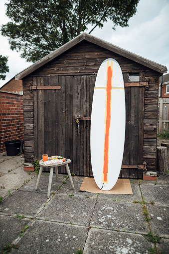 A wide shot of a surfboard leaning up against a shed in a back garden in Northeastern England. The surfboard has been partially painted down the middle.