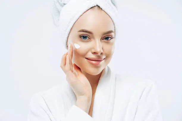 Photo of Cropped shot of pretty young woman applies cream for rejuvenation, healthy soft skin, uses cosmetic product, demonstrates nice effect of body lotion, wears comfortable soft white bath robe, towel