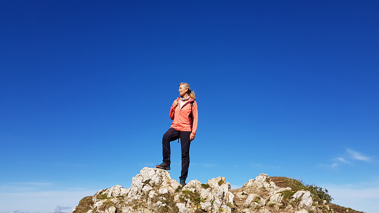 Active senior woman with blond hair carrying small backpack, hiking and enjoying in nature. Beautiful mountains and autumn trees in the background.