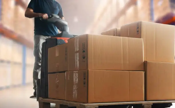 Photo of Shipment boxes, Cargo warehouse. Warehouse worker driving electric forklift pallet jack unloading pallet goods.