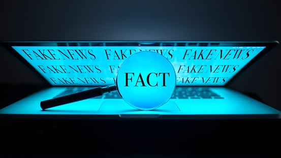 a magnifying glass on a laptop in the dark shows the word fact, in the background on the screen there are fake news