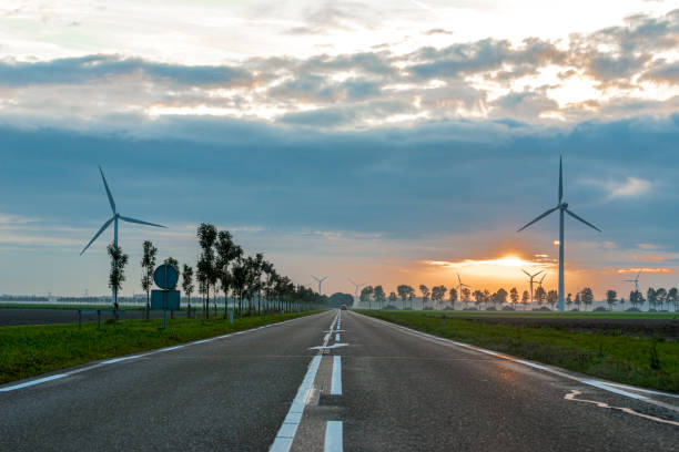 group of wind mills in the fog at sunset, in the foreground an empty road, setting sun reflecting on the road.