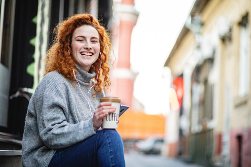 Portrait of a happy young woman in a city smiling and holding a smart phone and travel mug