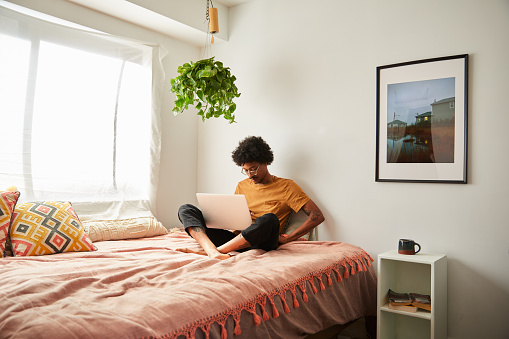 Young African American man sitting on his bed browsing online with a laptop and checking his cellphone