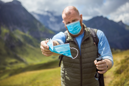 Male hiker man enjoying some free time and fresh air outdoors in the Alpine mountains on a beautiful day whilst wearing a protective corona virus face mask during the covid-19 outbreak