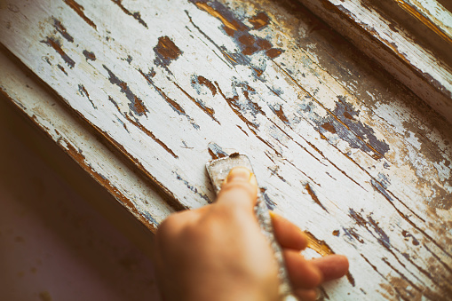 Female hand peels off an old layer of paint from a wooden window frame using spatula