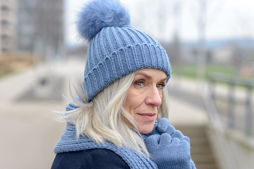 Pretty blond woman cuddling down into her warm knitted blue woolly scarf, hat and gloves as she stands waiting outdoors on a walkway in the city