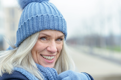 Happy blond woman wearing a warm knitted woollen beanie looking at the camera with a vivacious smile in a close up head shot outdoors in winter