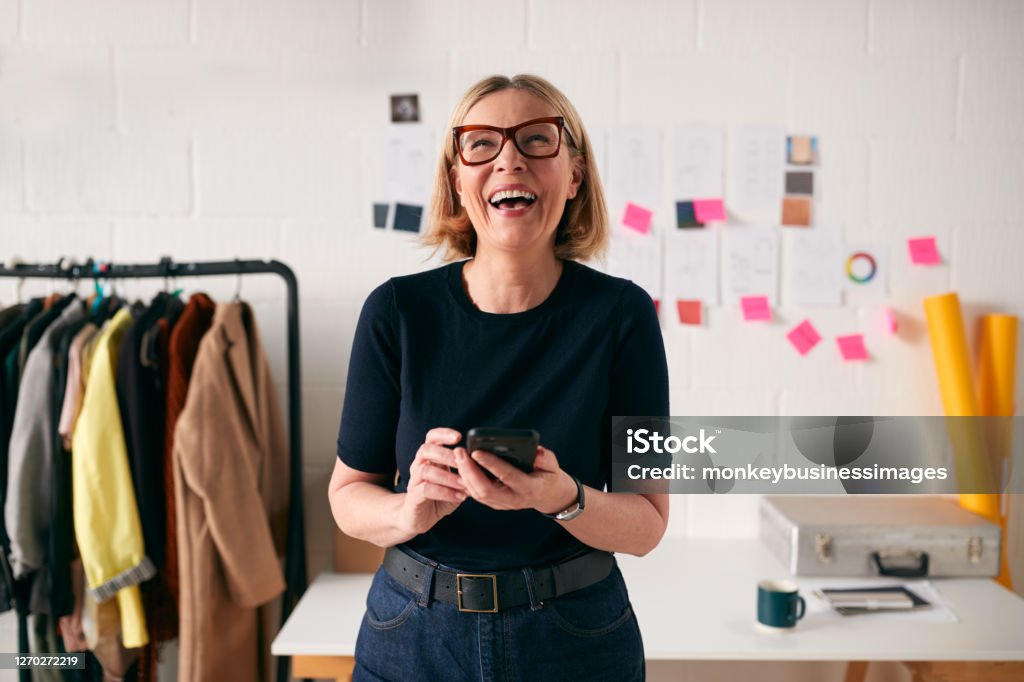 Laughing Mature Businesswoman With Mobile Phone In Front Of Desk In Start Up Fashion Business Women Stock Photo