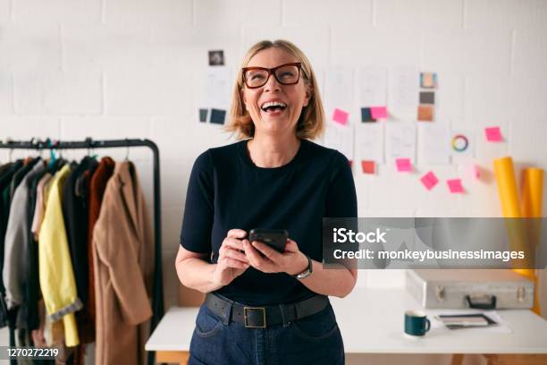 Laughing Mature Businesswoman With Mobile Phone In Front Of Desk In Start Up Fashion Business Stock Photo - Download Image Now