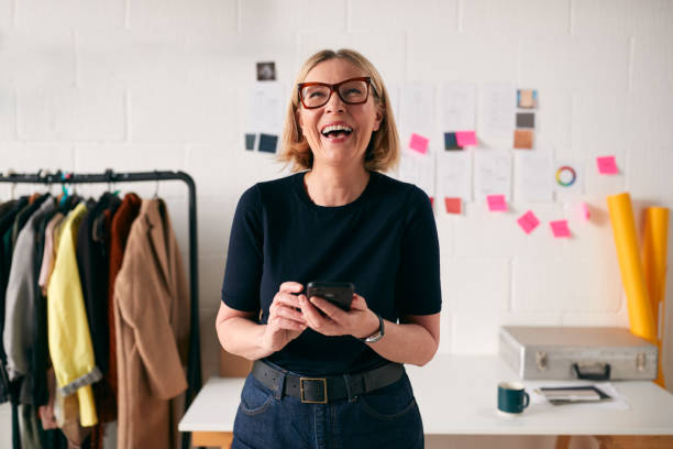 laughing mature businesswoman with mobile phone in front of desk in start up fashion business - business owner imagens e fotografias de stock