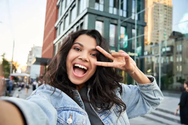 Happy African American hipster teen girl blogger influencer recording vlog holding looking at camera. Black woman travel vlogger influencer shooting video, streaming on urban city street, headshot.