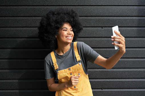 Happy African American hipster teen girl blogger with Afro hair laughing, holding phone, recording vlog, shooting social media stories or video calling in mobile app standing on black background.