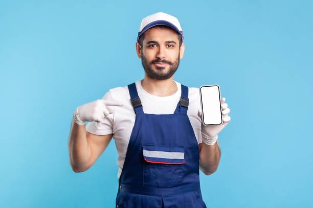 istock Workman in overalls and gloves pointing mobile phone with mock up blank display, advertising area 1270275521