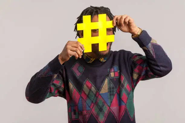 Photo of Playful happy african guy with dreadlocks in trendy sweatshirt hiding face behind hashtag symbol, popular blogger recommending to follow tag, comments