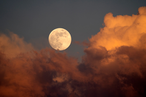 A fell moon rises from behind the sunset-lit clouds.