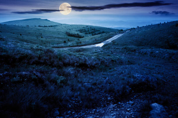 mountain road through grassy meadow at night mountain road through grassy meadow at night. wonderful summer adventure in full moon light. clouds on the blue sky country road sky field cloudscape stock pictures, royalty-free photos & images