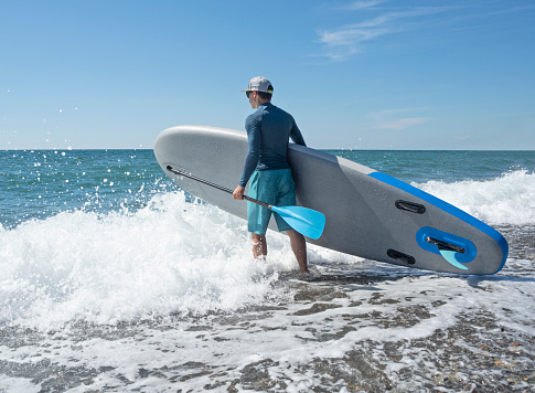 Young active person with inflatable stand up paddle board and oar in sea waves go surfing