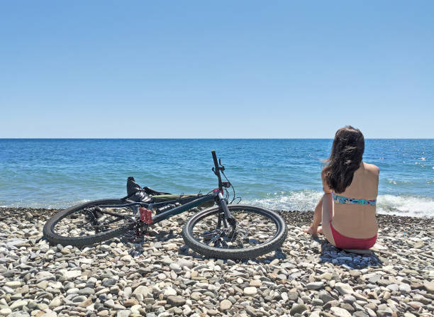Back view of active girl sitting on sea coast with bicycle and relaxing after bike trip