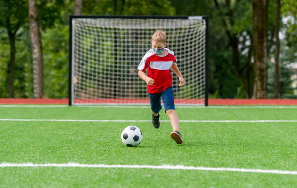 ragazzo della scuola con maschera e pallone da calcio in una lezione di educazione fisica. ritorno a scuola durante il concetto di pandemia. distanziamento sociale per combattere il covid-19 - little boys people indoors soccer foto e immagini stock