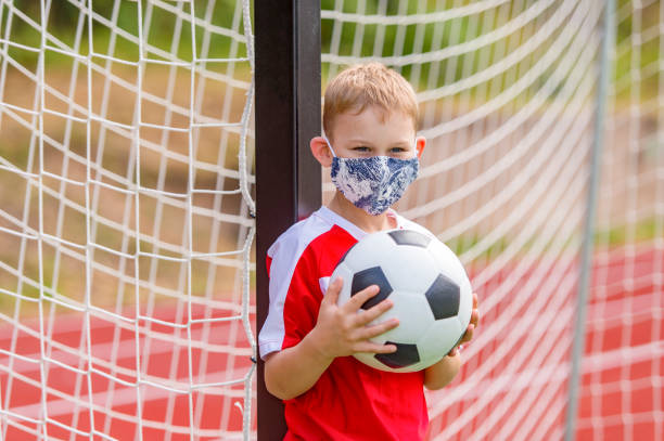 School kid with mask and soccer ball in a physical education lesson. Safe back to school during pandemic concept. Social distancing to fight COVID-19 School kid with mask and soccer ball in a physical education lesson. Safe back to school during pandemic concept. Social distancing to fight COVID-19 face guard sport photos stock pictures, royalty-free photos & images