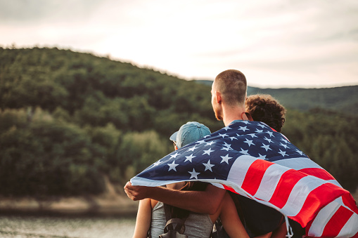 Small caucasian group of young people walking in nature near lake and forest and proudly showing American flag.