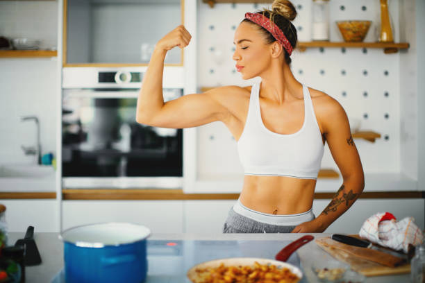 Nutrition is just as important as exercise. Young fit woman is preparing a protein meal in the kitchen. She standing and proudly showing her muscular hand. ketogenic diet stock pictures, royalty-free photos & images