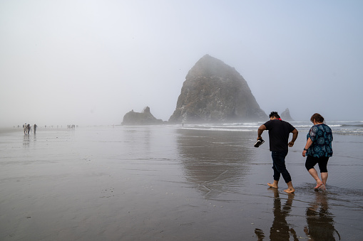 Cannon Beach, Oregon - August 1, 2020: Tourist couple walks barefoot in the ocean near Haystack Rock on a foggy morning