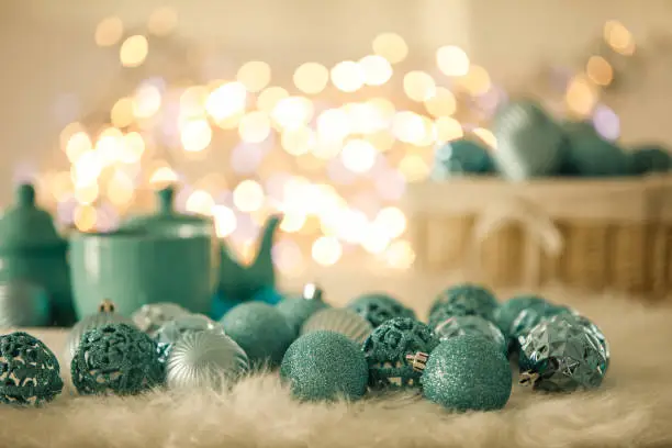 Copy space shot of arrangement of bunch of turquoise colored Christmas bobbles on a white faux fur rug with some tea pots, tea cups and blurred fairy lights in the background.