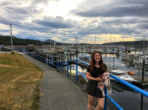 A pretty young female tourist standing at the waterfront in Port McNeil with the marina in the background, on a pretty evening on Northern Vancouver Island, British Columbia, Canada