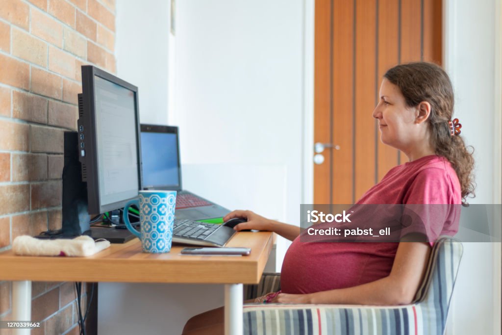 Pregnant woman smiling working from home living room Pregnant woman sitting in front of her work desk, working on the computer. Working Class Stock Photo