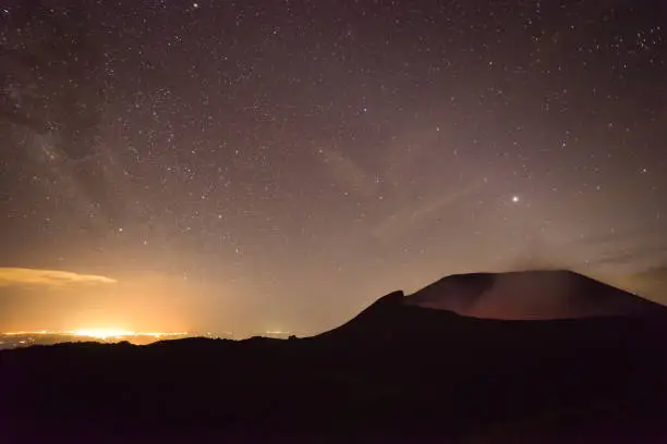 Photo of scenic panorama of the active volcano Telica with sulfuric smoke coming out of the volcanic cone at night with starry sky and lights of the city of León in the background