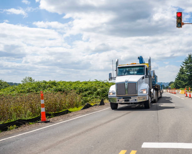 Big rig semi truck transporting heavy equipment on step down semi trailer moves along a section of road with road works White big rig industrial diesel day cab semi truck with semi trailer standing on the temporary installed mobile traffic light stop point on the repairing section of the road in Columbia Gorge shipping heavy equipment machinery stock pictures, royalty-free photos & images
