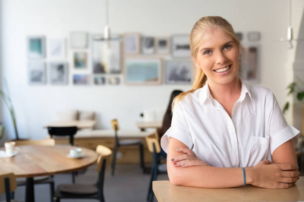 Happy beautiful blonde woman wearing white shirt Happy beautiful blonde woman wearing white shirt, standing in co-working space, leaning on desk, posing, looking at camera and smiling. Medium shot, copy space. People at co-working concept medium shot stock pictures, royalty-free photos & images